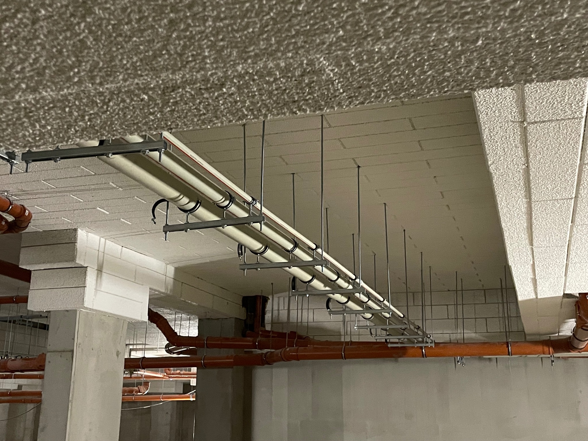 Instalacja rur pod sufitem w surowym stanie. Rury kanalizacyjne w kolorze pomarańczowym i białe rury wodociągowe zamontowane na metalowych wspornikach.