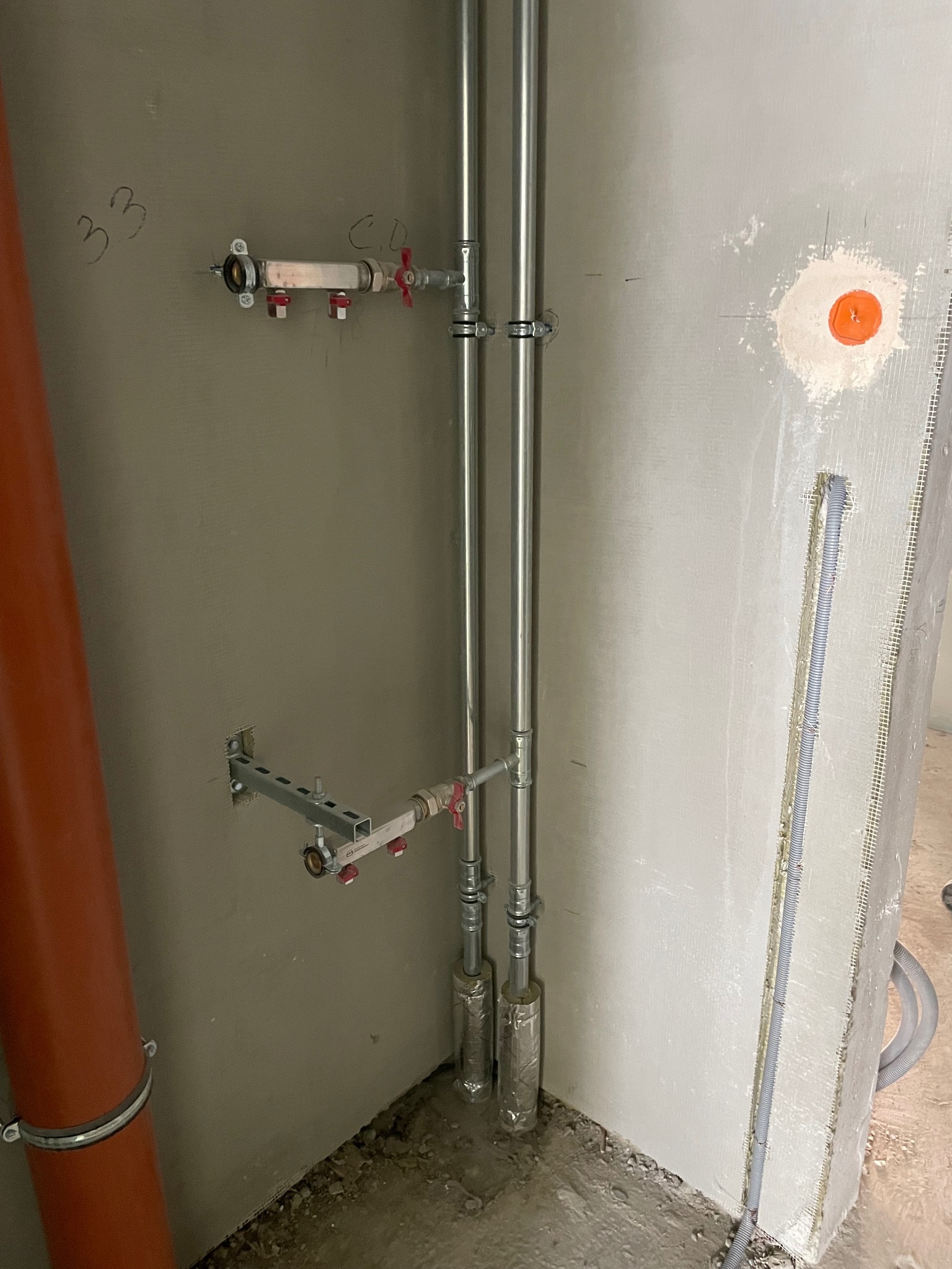 Szare ściany z instalacją: rury pionowe, poziome z zaworami, mocowane do ściany na wspornikach. Pomarańczowa rura kanalizacyjna w rogu. Widoczne ślady po pracach instalacyjnych.