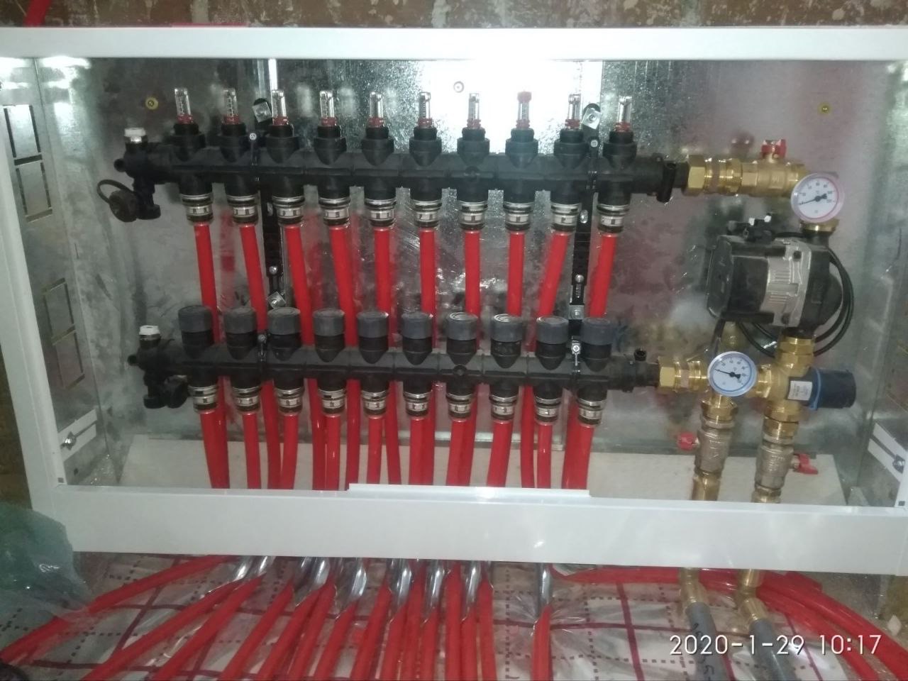 Rozdzielacz ogrzewania podłogowego w metalowej szafce, z czerwonymi rurami, manometrami i pompą obiegową. Widoczne datowniki na podłodze.