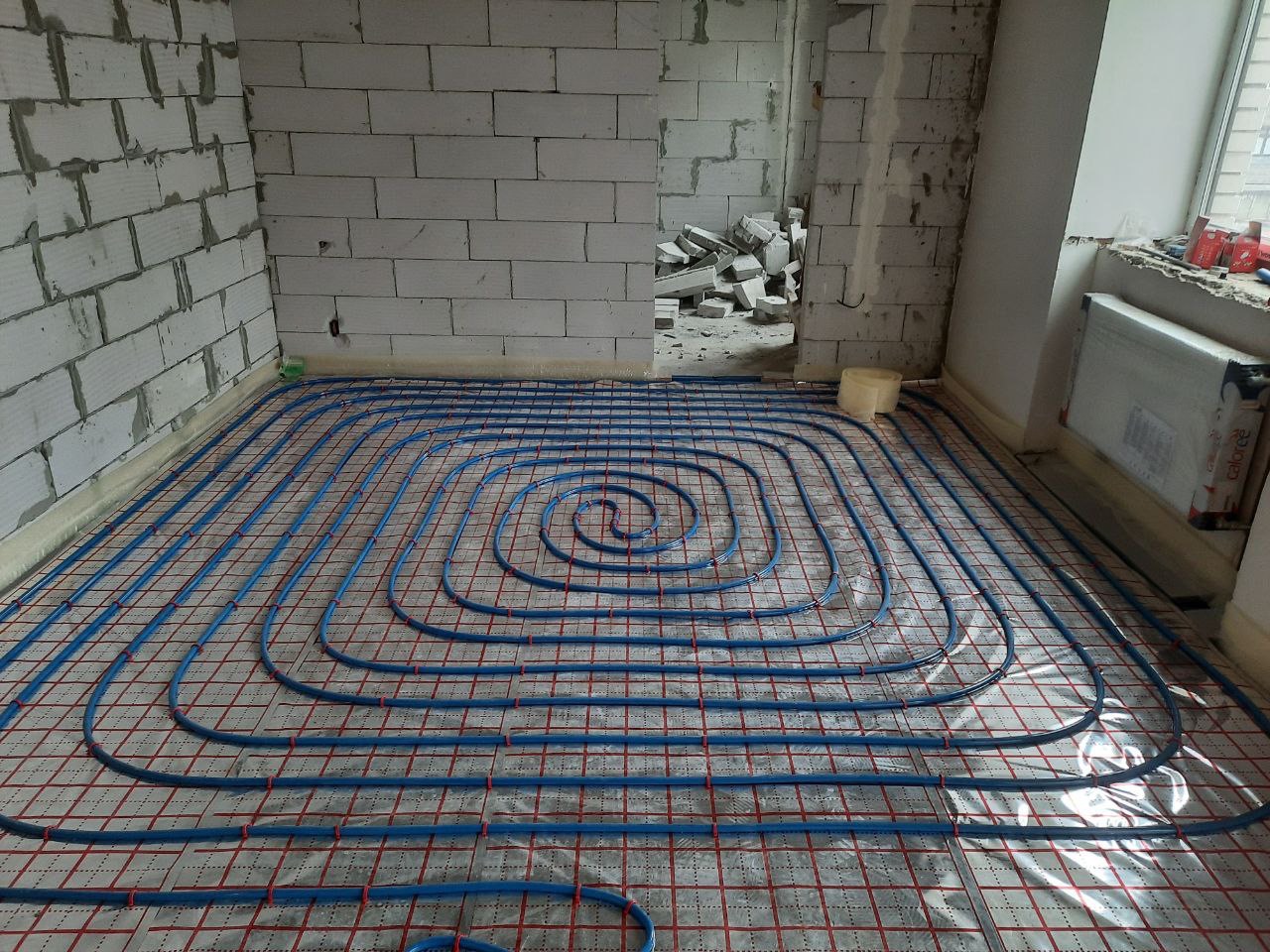 Niebieskie rury ogrzewania podłogowego ułożone spiralnie na czerwonej siatce w surowym wnętrzu z białego bloczka, widok z góry, etap instalacji przed wylewką.
