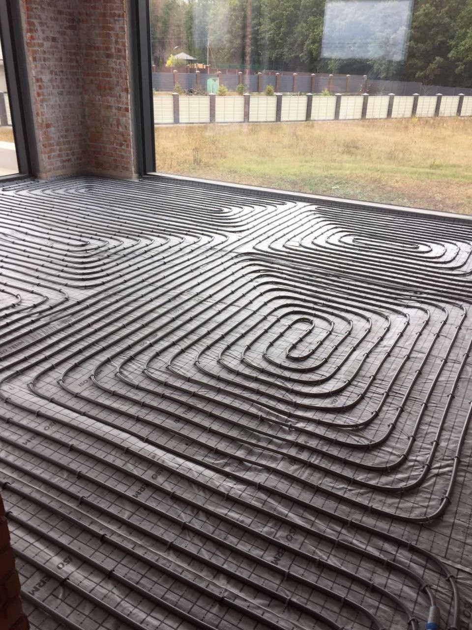 Ułożona instalacja ogrzewania podłogowego w pokoju z dużym oknem i ceglanym filarem. Widoczne rury grzewcze na folii izolacyjnej przed wylaniem posadzki.