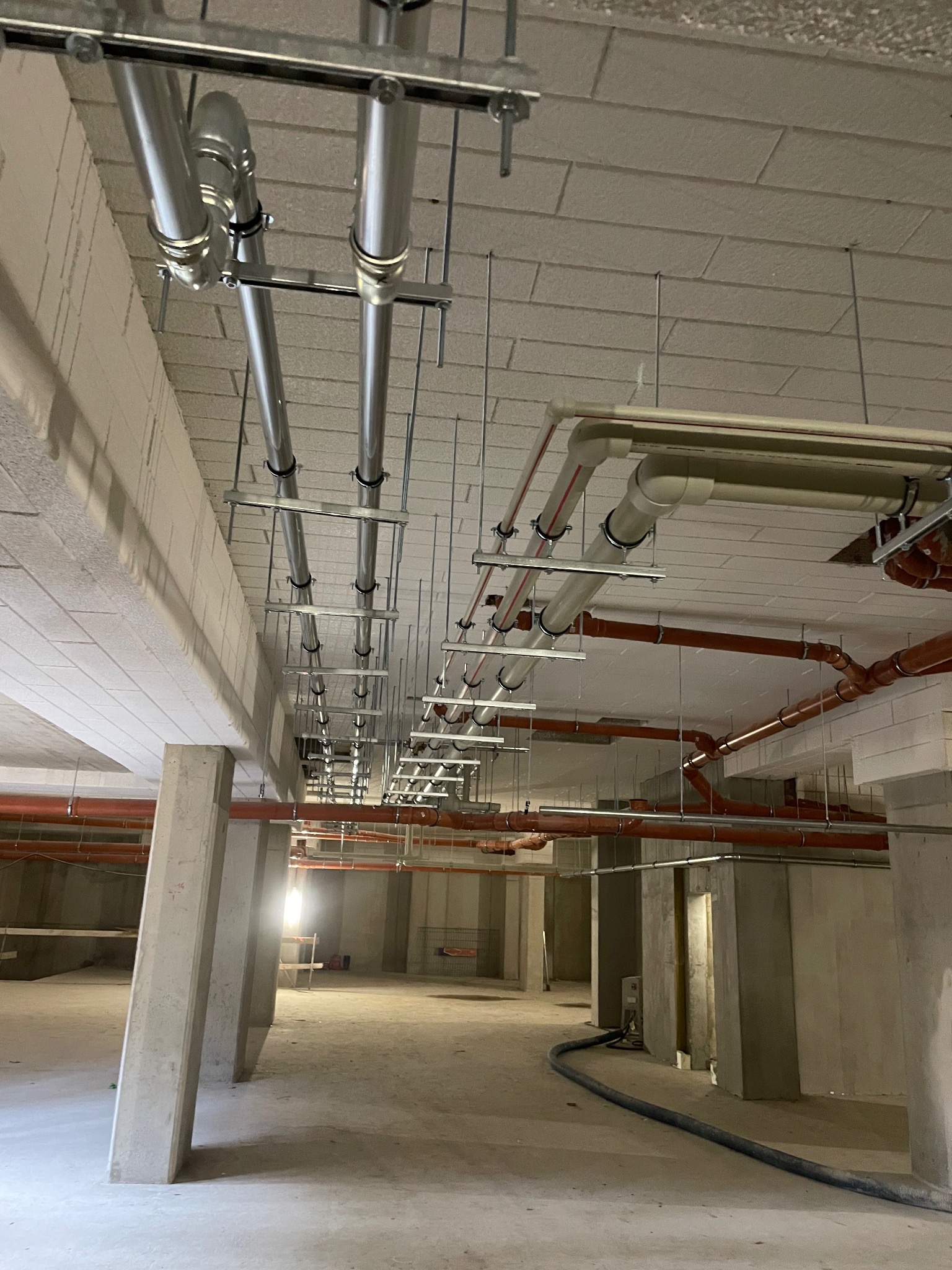 Widok instalacji pod stropem: rury stalowe i PCV, podwieszone na metalowych konstrukcjach. Stan surowy, brak wykończenia wnętrz. Widoczna perspektywa długiego korytarza.