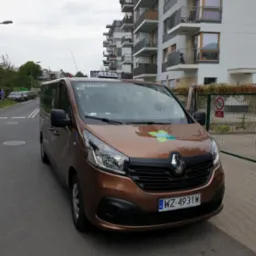 Brązowy van Renault Trafic z otwartymi drzwiami na tle bloku mieszkalnego. Na masce logo EKO Taxi, na dachu napis TAXI. Tablica rejestracyjna WZ 4931W.