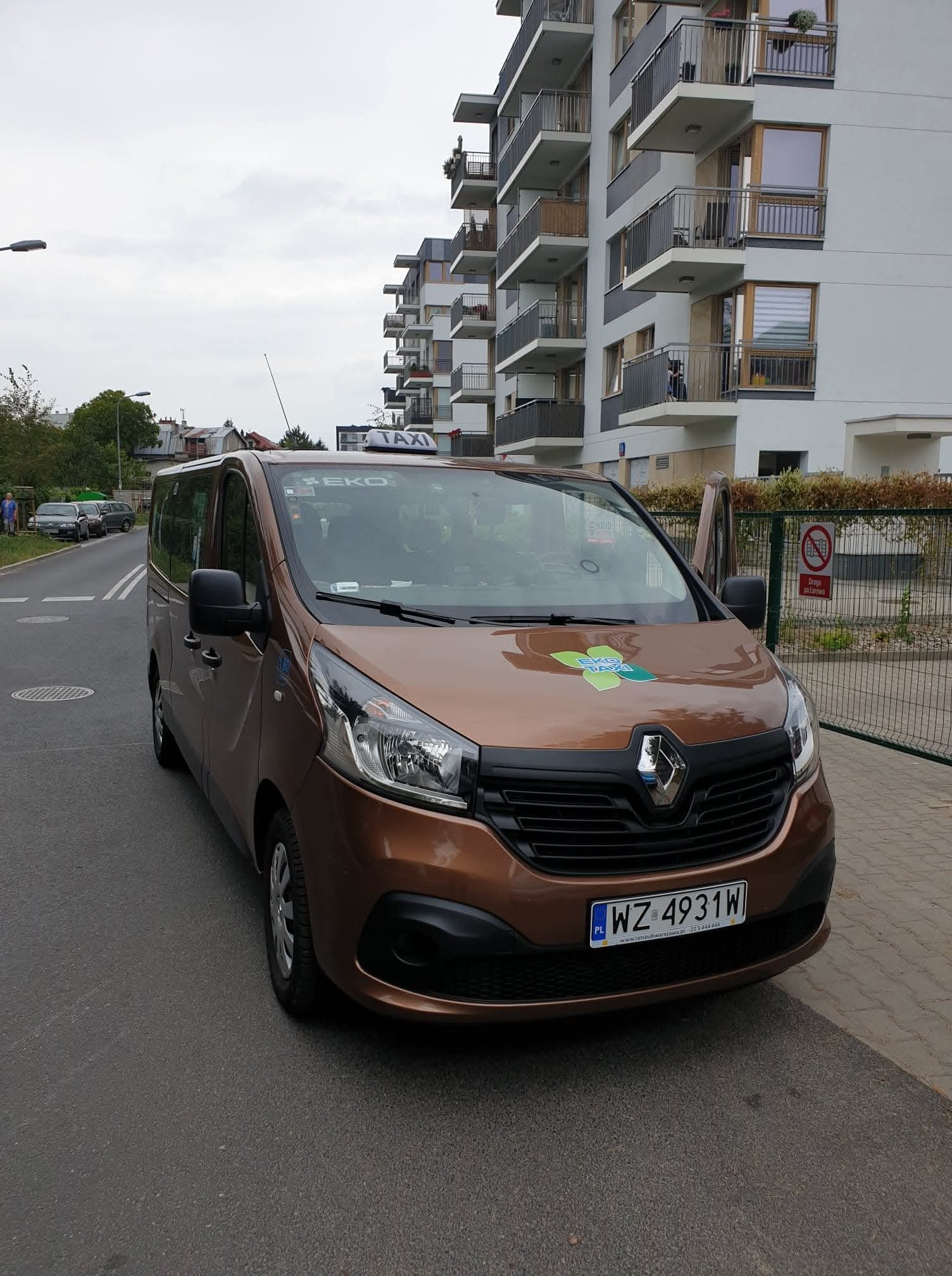 Brązowy van Renault Trafic z otwartymi drzwiami na tle bloku mieszkalnego. Na masce logo EKO Taxi, na dachu napis TAXI. Tablica rejestracyjna WZ 4931W.