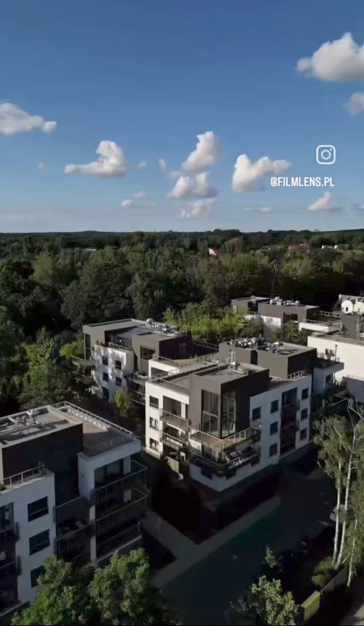 Nowoczesne osiedle z lotu ptaka w słoneczny dzień. Białe budynki z szarymi akcentami, balkony z meblami, zielone drzewa i błękitne niebo z chmurami. Logo w rogu.