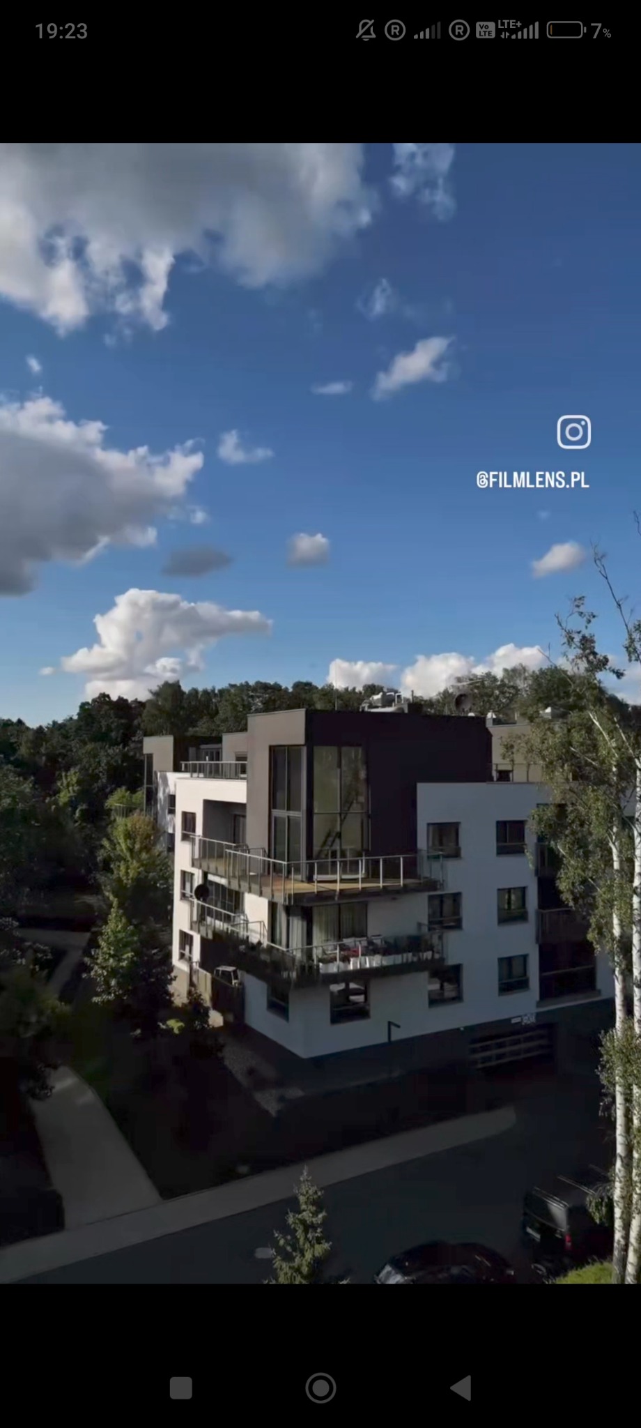 Nowoczesny budynek wielorodzinny z balkonami, sfotografowany z góry w słoneczny dzień z logiem @filmlens.pl. Widoczne drzewa i zaparkowane samochody.