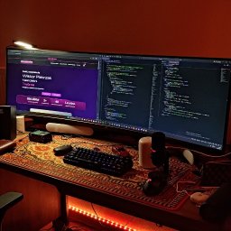 Web Nest - Stanowisko programisty: dwa monitory, na jednym kod, na drugim strona internetowa. Biurko z wzorzystą podkładką, klawiatura, myszka, mikrofon. Czerwone oświetlenie LED.