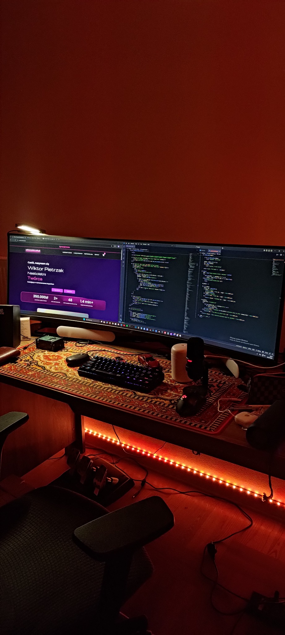 Stanowisko programisty: dwa monitory, na jednym kod, na drugim strona internetowa. Biurko z wzorzystą podkładką, klawiatura, myszka, mikrofon. Czerwone oświetlenie LED.