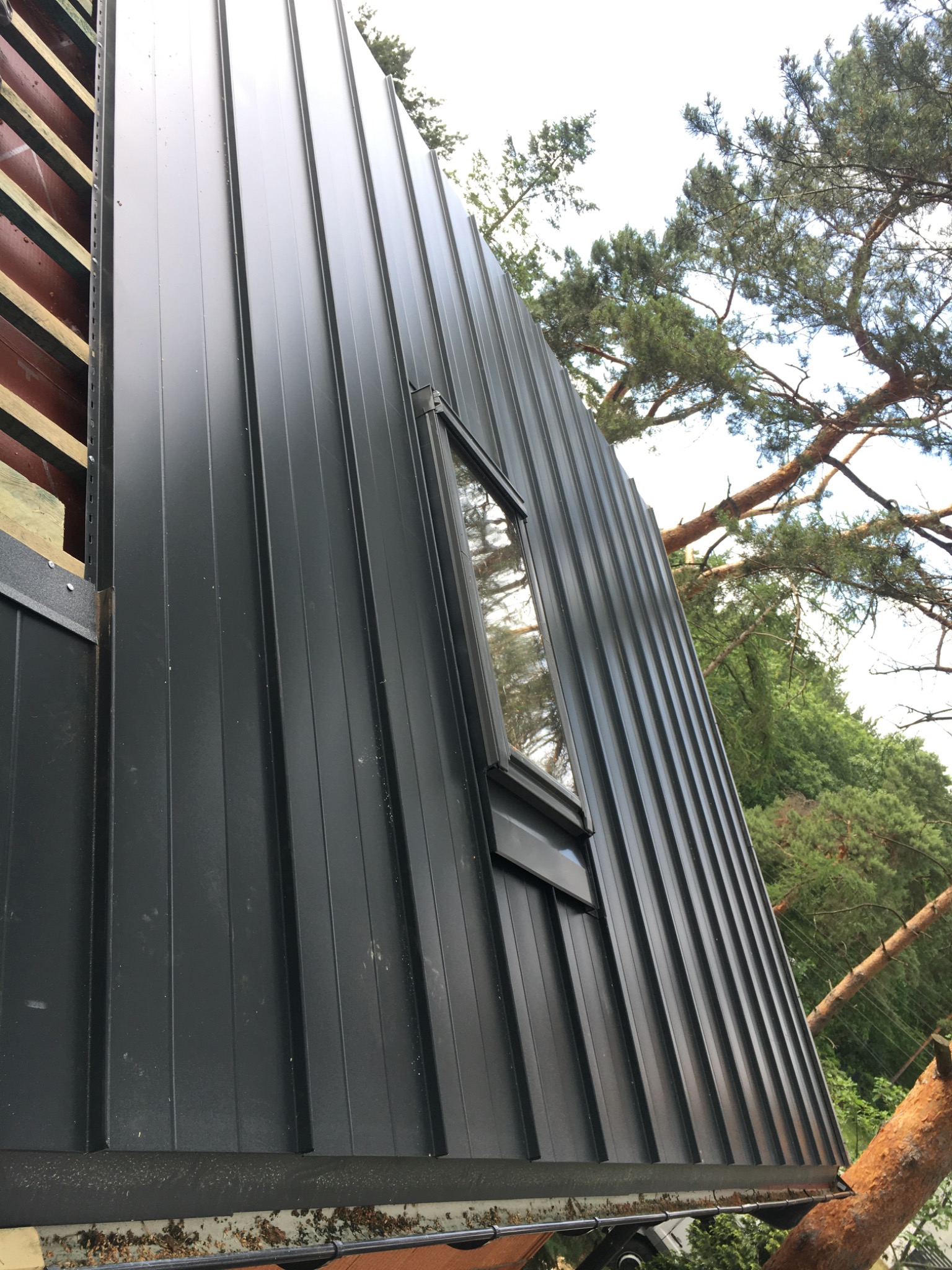 Nowoczesne krycie dachu blachą na rąbek stojący w kolorze grafitowym, z oknem dachowym. Widok z dołu, na tle drzew.