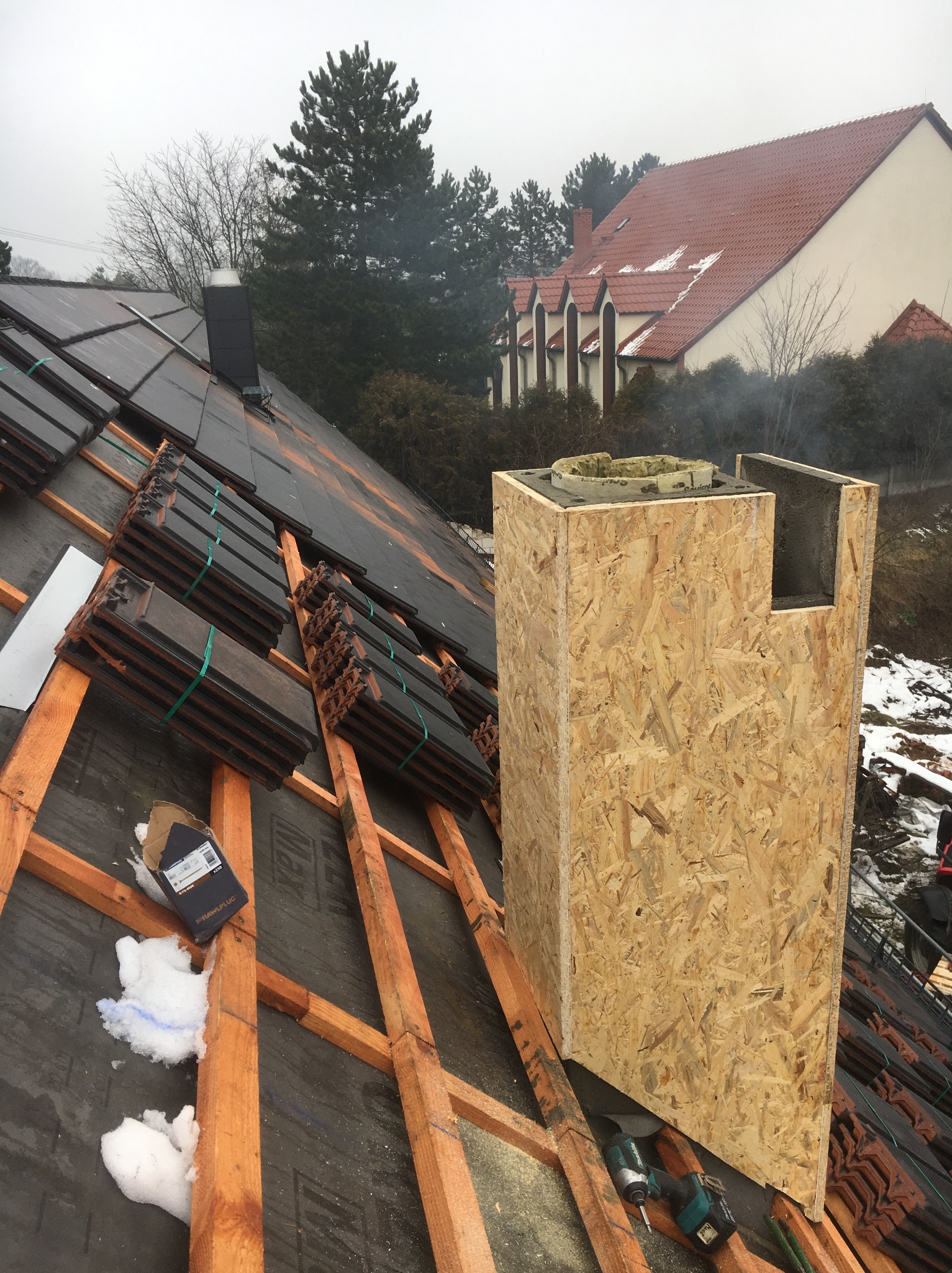 Montaż komina z osb na dachu pokrytym papą i częściowo dachówką. Widoczne narzędzia i elementy konstrukcji dachu. W tle budynek i drzewa.