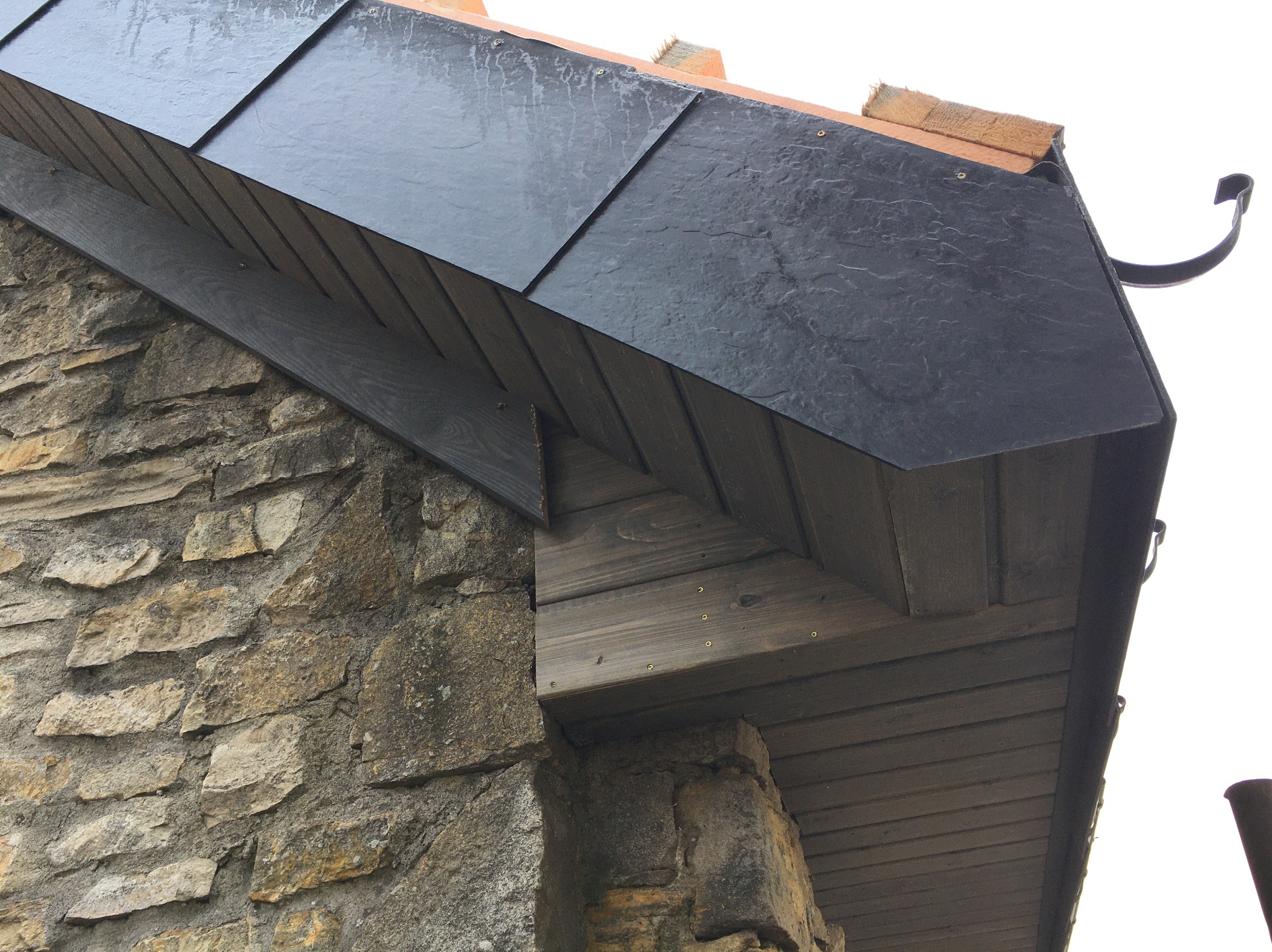 Fragment dachu krytego czarnym łupkiem, z widoczną drewnianą konstrukcją okapu i kamienną ścianą budynku. Ujęcie z dołu, podkreślające detale wykończenia.