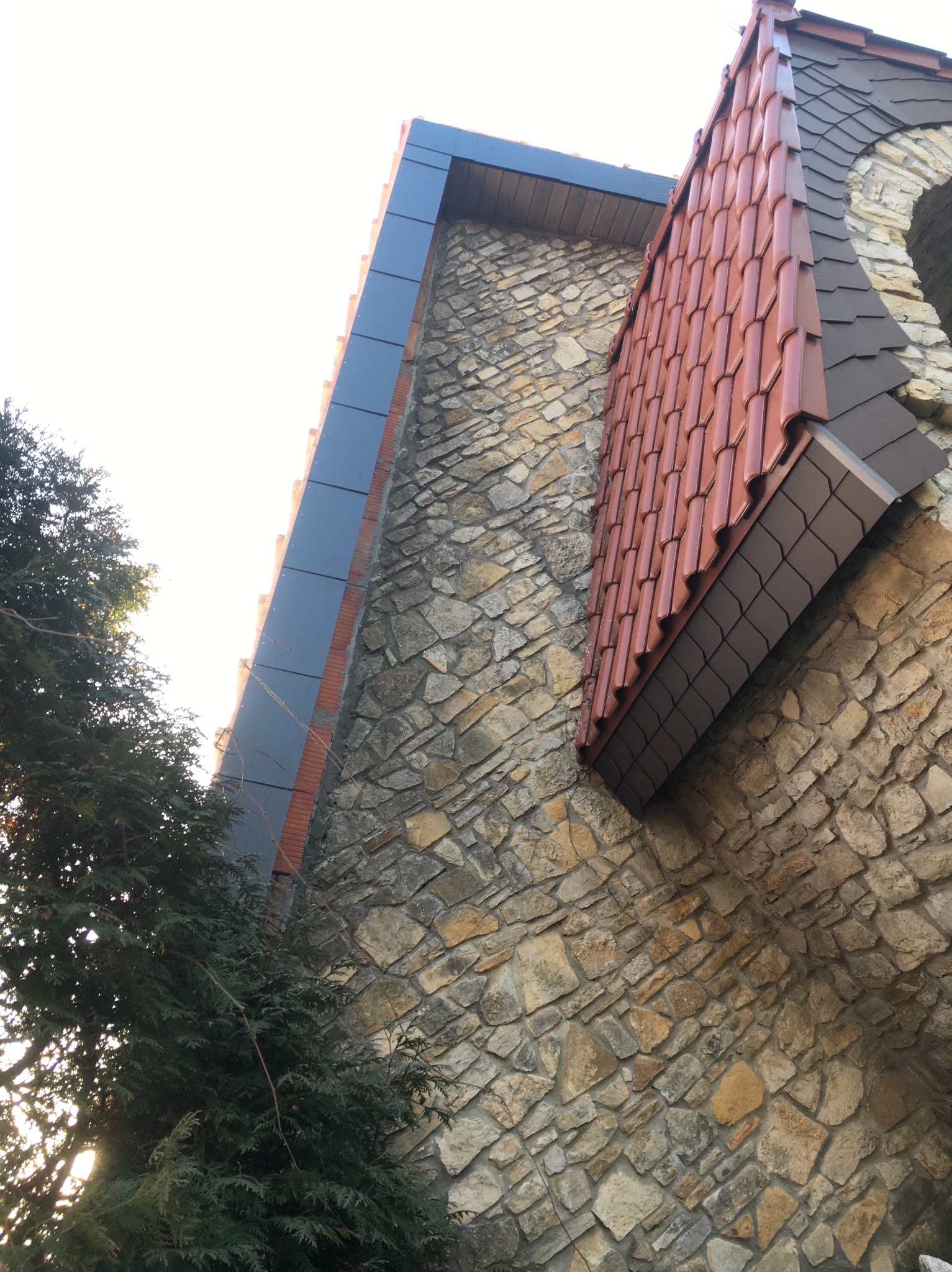 Fragment elewacji budynku w Warszawie z kamienia, z nowym kryciem dachu z czerwonej dachówki i obróbkami blacharskimi w kolorze grafitowym.