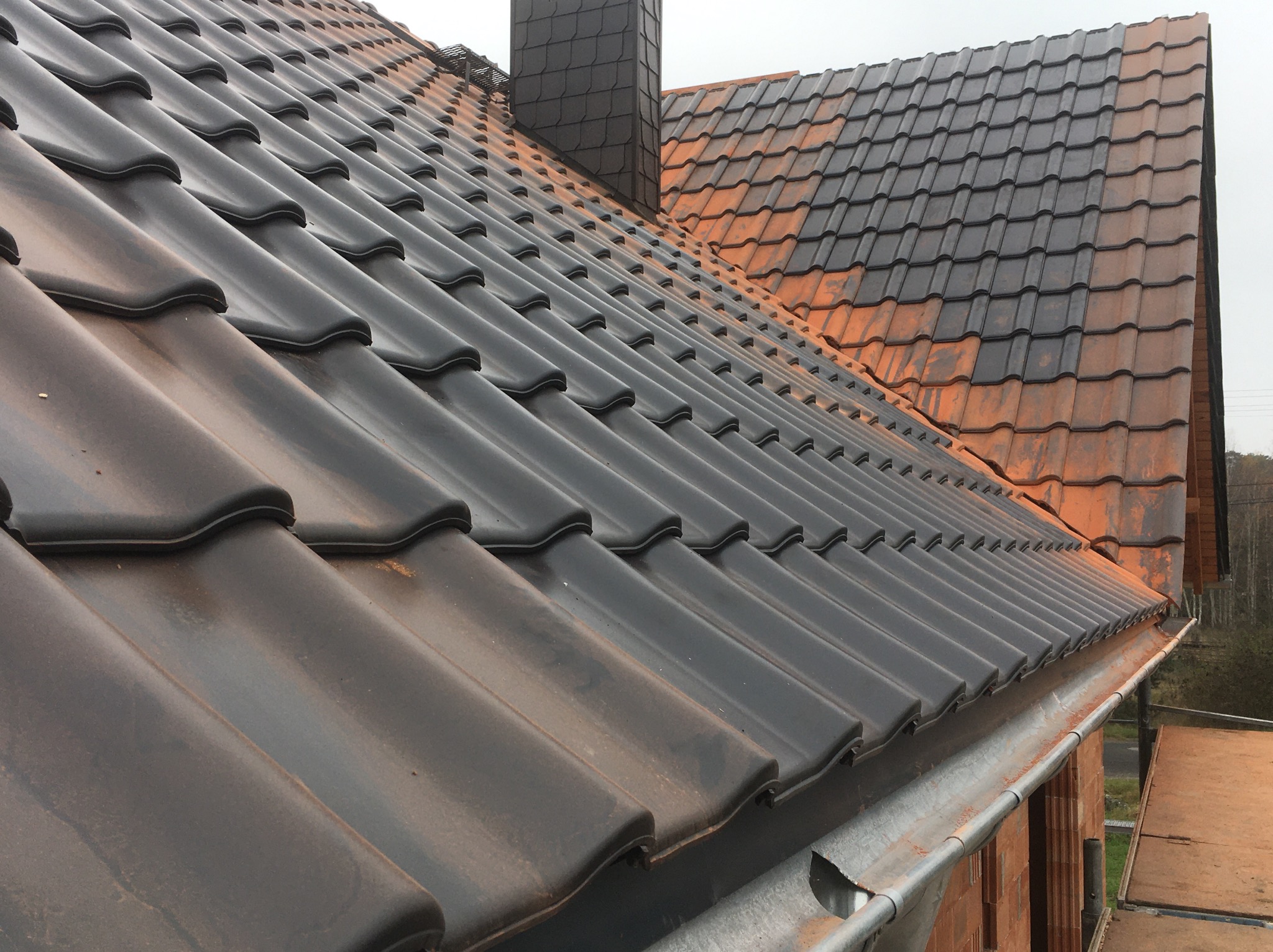 Ciemna dachówka ceramiczna na skośnym dachu z widocznym kominem i rynną. Część dachu pokryta starą, pomarańczową dachówką. Widok z góry na fragment dachu.