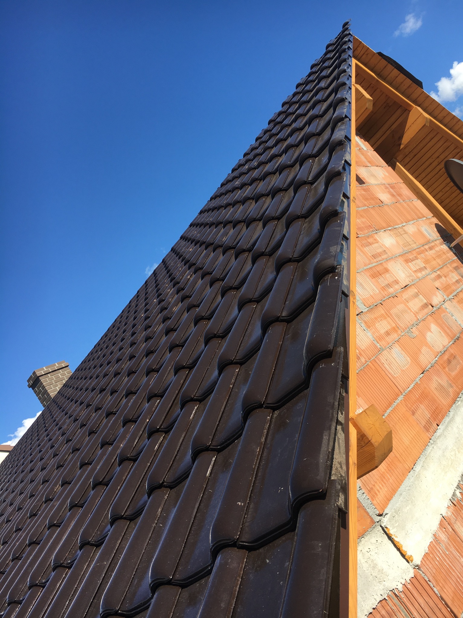 Ciemna dachówka ceramiczna na skośnym dachu, widok z dołu, z fragmentem muru z pustaków ceramicznych i drewnianą konstrukcją więźby dachowej na tle błękitnego nieba.