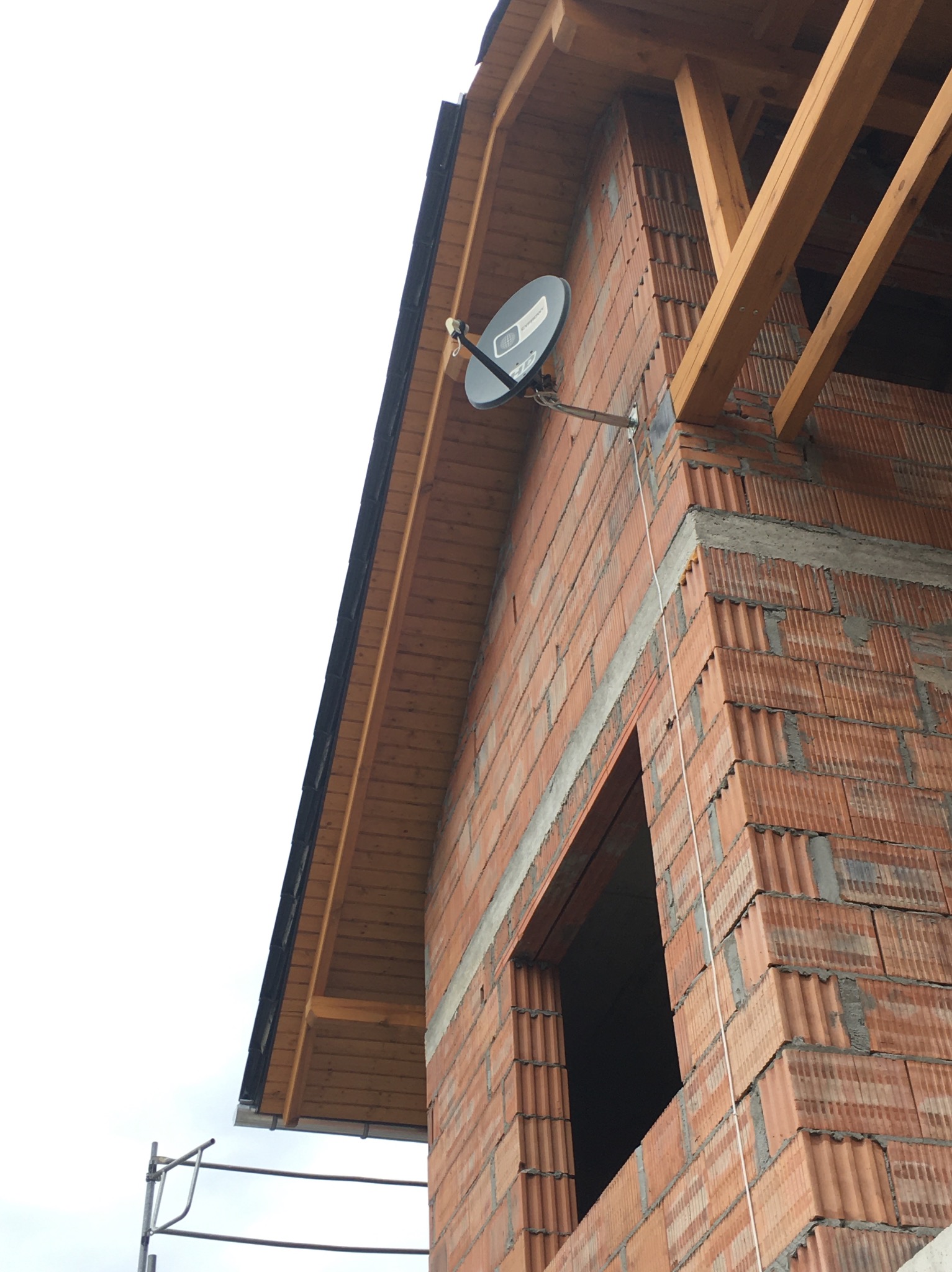 Fragment ściany z czerwonej cegły z oknem w stanie surowym, widoczny fragment dachu z drewnianą podbitką i zamontowana antena satelitarna na elewacji budynku.