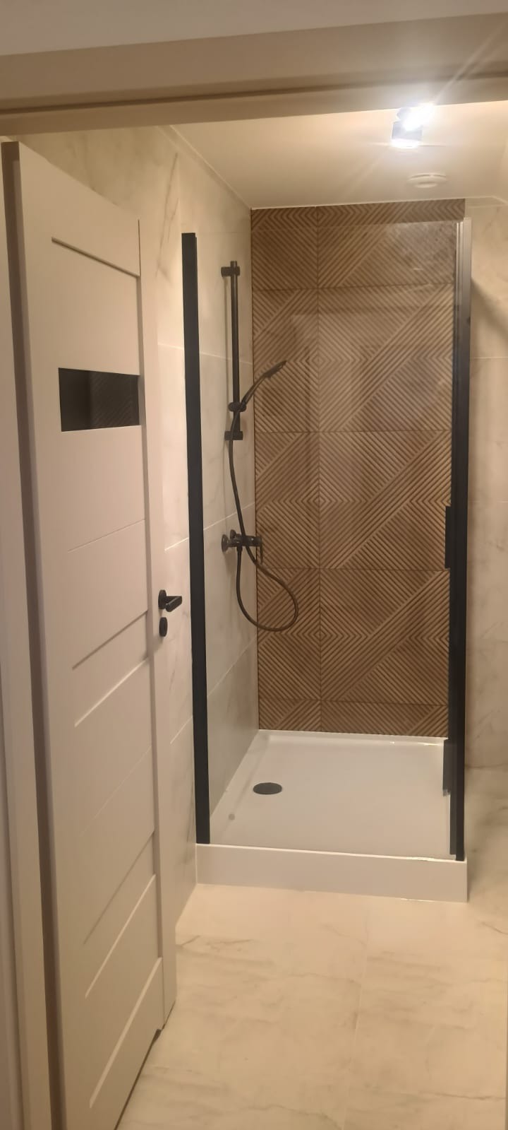 Nowoczesna łazienka po remoncie: kabina prysznicowa z czarnymi profilami, deszczownica i dekoracyjna ściana z płytek o geometrycznym wzorze. Białe drzwi z czarnym akcentem.