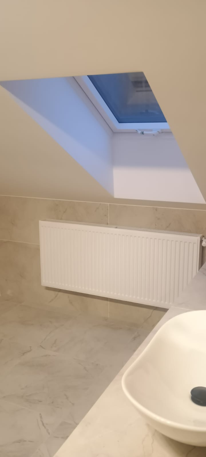 Jasna łazienka na poddaszu z oknem dachowym. Biały grzejnik na ścianie z płytek imitujących marmur. Umywalka nablatowa z czarnym korkiem. Minimalistyczny design.