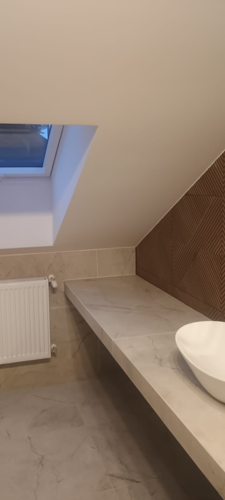 Wykończona łazienka na poddaszu w Wyszatycach: marmurowe płytki, blat pod umywalkę i dekoracyjna ściana. Okno dachowe wpuszcza naturalne światło.