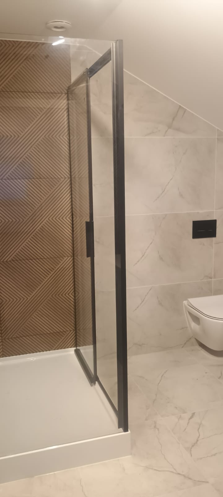 Nowoczesna kabina prysznicowa z czarnymi profilami, drewnopodobna ściana z geometrycznym wzorem, marmurowe płytki i podwieszana toaleta w wykończonej łazience.