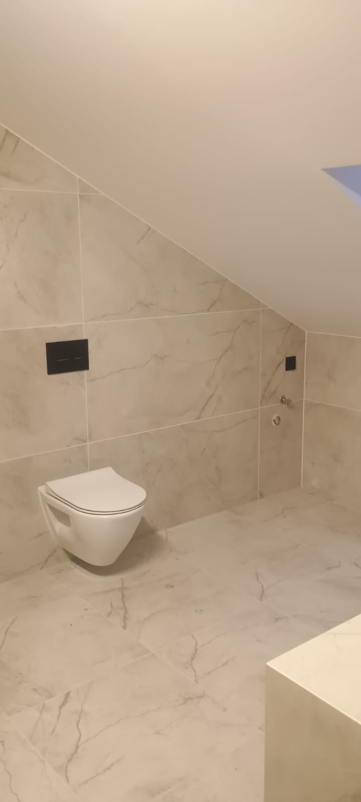 Wykończona łazienka na poddaszu ze ścianami i podłogą wyłożonymi płytkami imitującymi marmur. Wisząca toaleta z czarnym przyciskiem spłukującym.