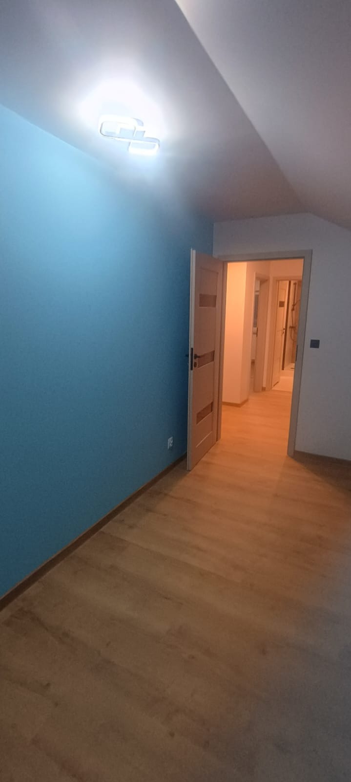 Błękitna ściana w pokoju z drewnianą podłogą i otwartymi drzwiami. Widoczna lampa sufitowa i fragment skosu dachu. Perspektywa wnętrza z widokiem na kolejne pomieszczenia.