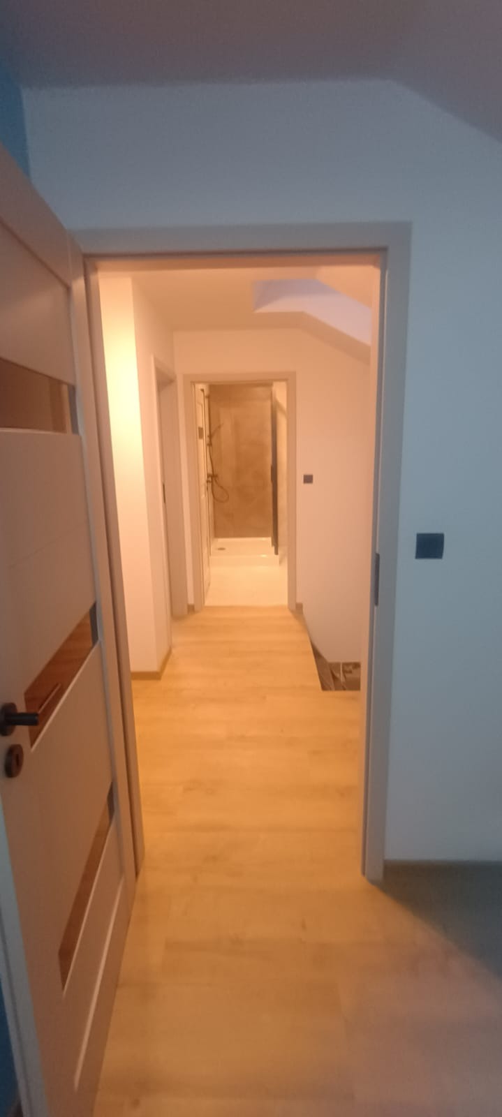 Jasne wnętrze po remoncie: widok na korytarz z drewnianą podłogą i otwarte drzwi do łazienki z kabiną prysznicową. Minimalistyczny design, świeżo po wykończeniu.