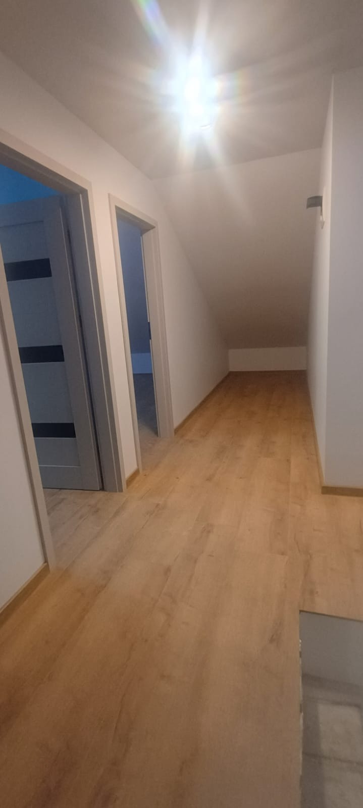 Jasne, wykończone poddasze z drewnianą podłogą i dwoma otwartymi drzwiami. Widoczne skosy i oświetlenie sufitowe. Minimalistyczny styl wykończenia.