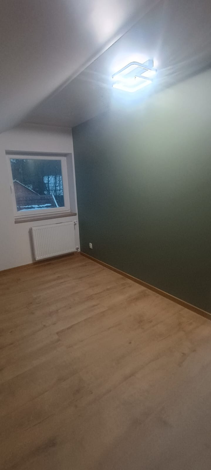 Pomalowane na oliwkowo wnętrze pokoju na poddaszu z drewnianą podłogą, oświetlone nowoczesną lampą sufitową. Widoczne okno z grzejnikiem.