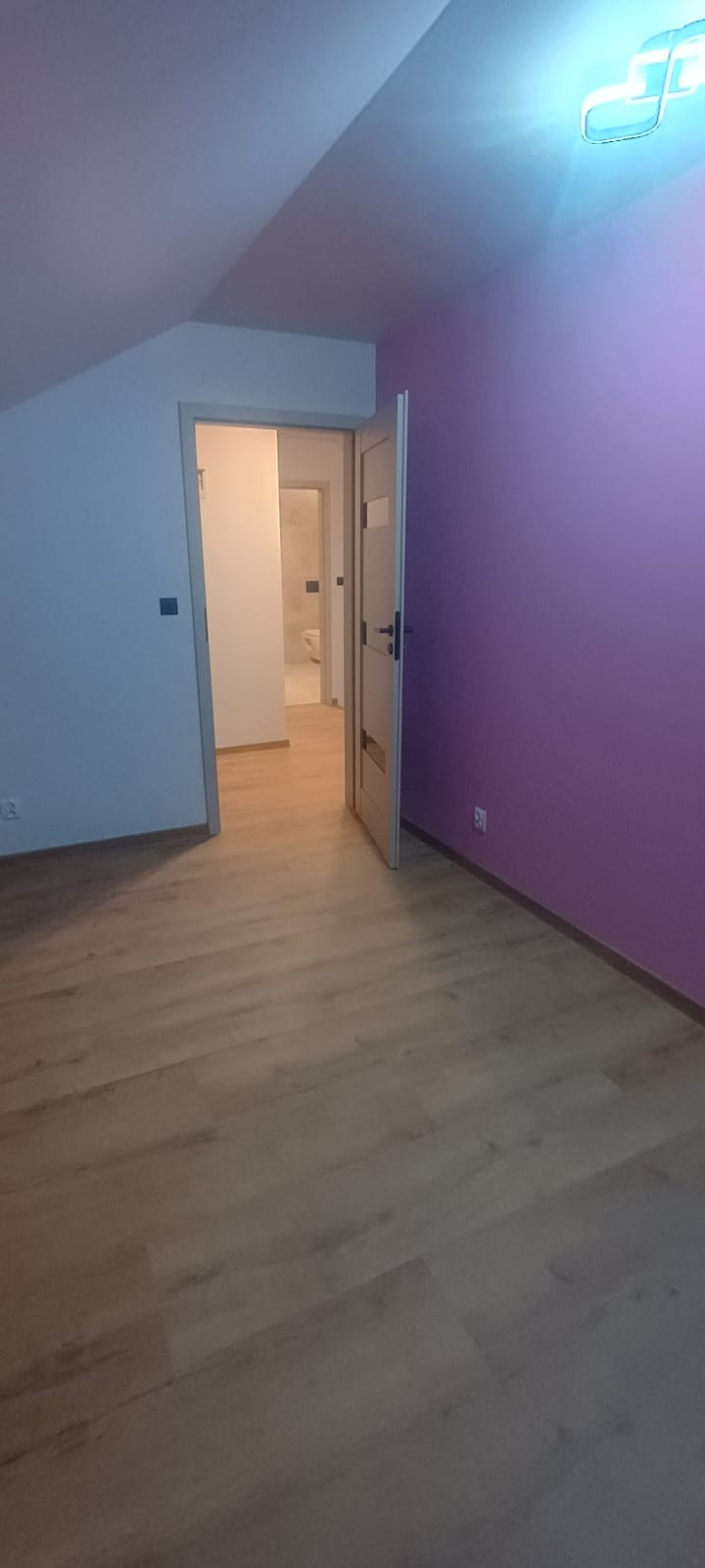 Jasne panele podłogowe w pokoju z fioletową ścianą i otwartymi, drewnianymi drzwiami. W tle widoczna łazienka z toaletą. Skos sufitu.