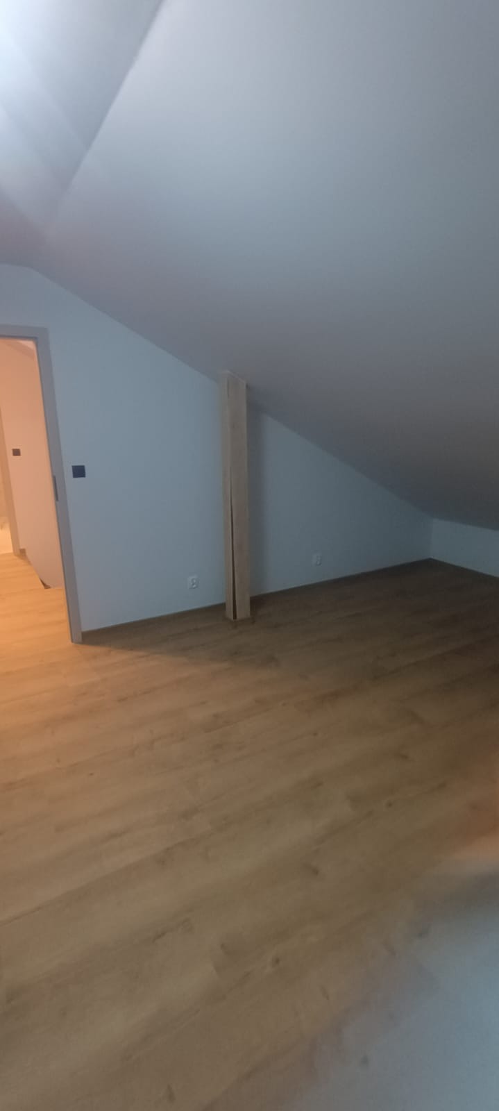 Jasne panele podłogowe w pokoju na poddaszu z widoczną drewnianą konstrukcją wsporczą i świeżo pomalowanymi ścianami. Widok na otwarte drzwi do innego pomieszczenia.