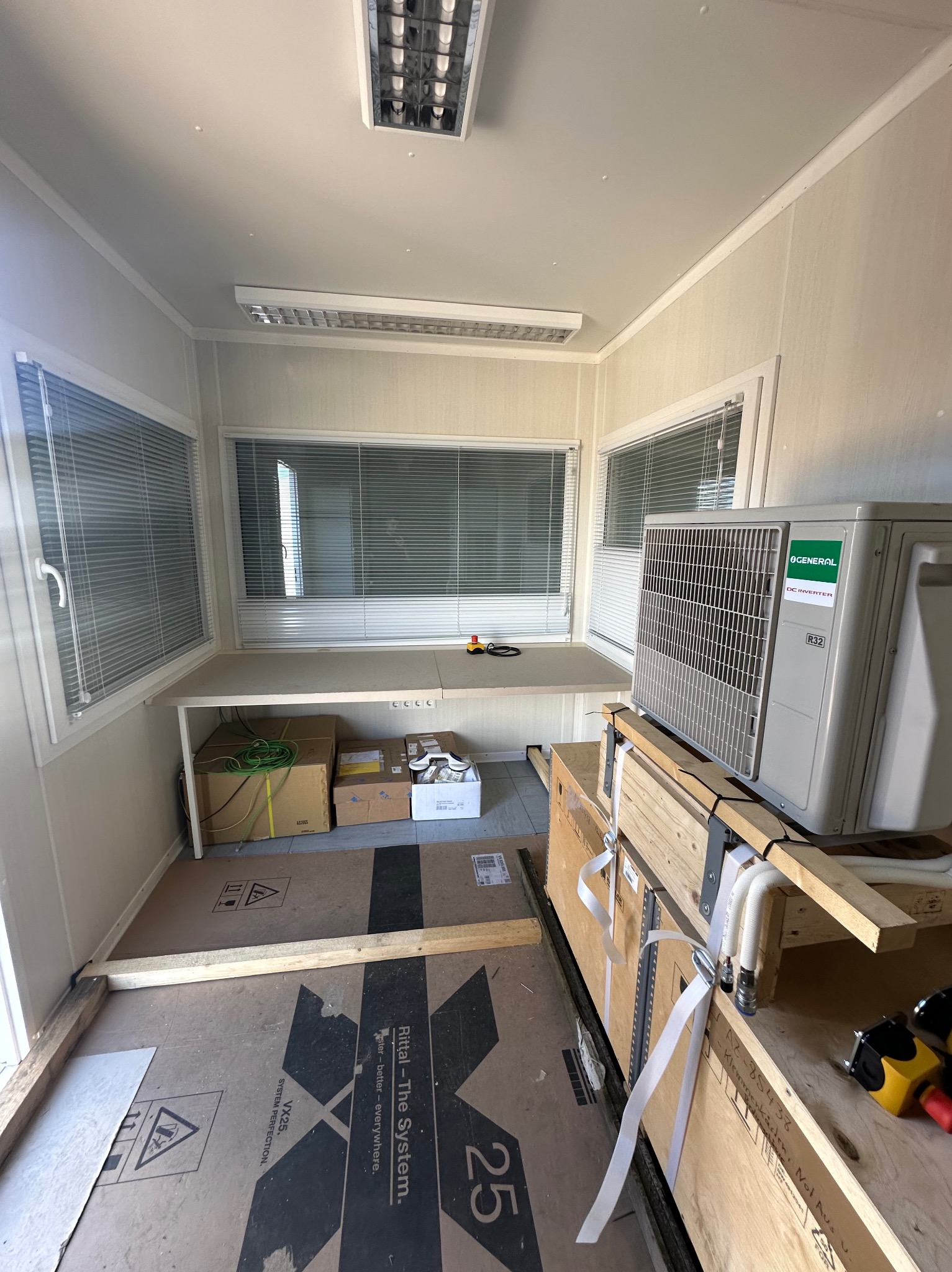 Wnętrze kontenera biurowego z oknami w żaluzjach i zamontowanym klimatyzatorem. Widoczne kable, skrzynki i narzędzia, sugerujące etap instalacji lub przygotowania.