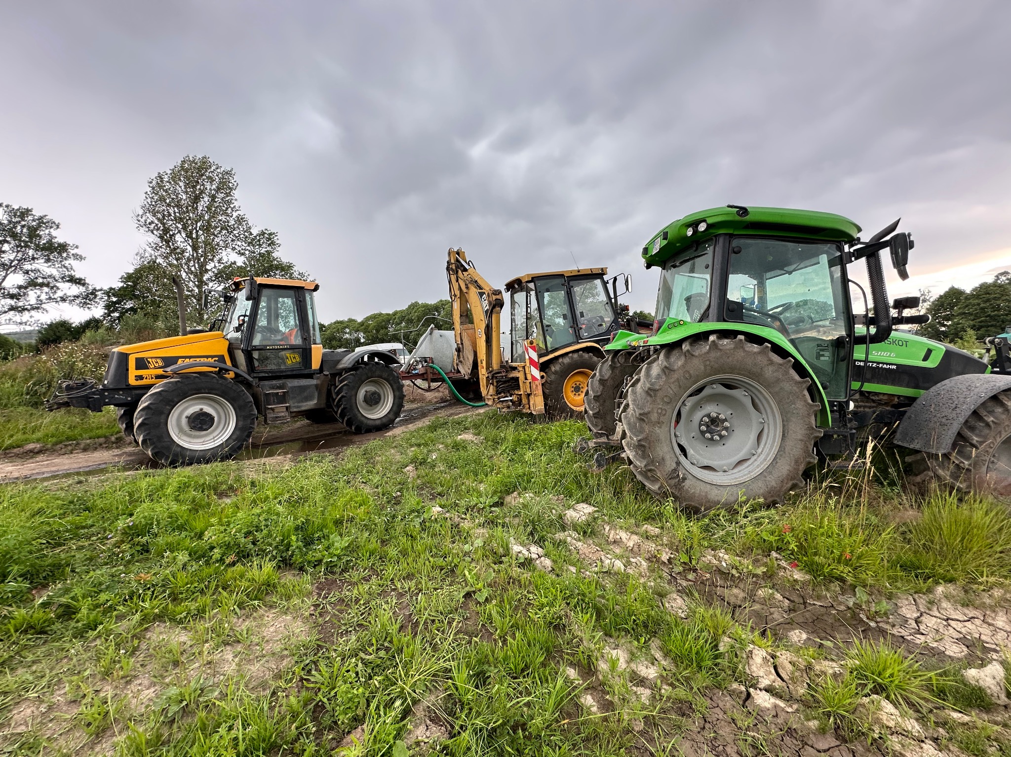 Żółty ciągnik JCB Fastrac, koparko-ładowarka i zielony ciągnik Deutz-Fahr na polu z zielenią, pod pochmurnym niebem. Sprzęt gotowy do pracy.