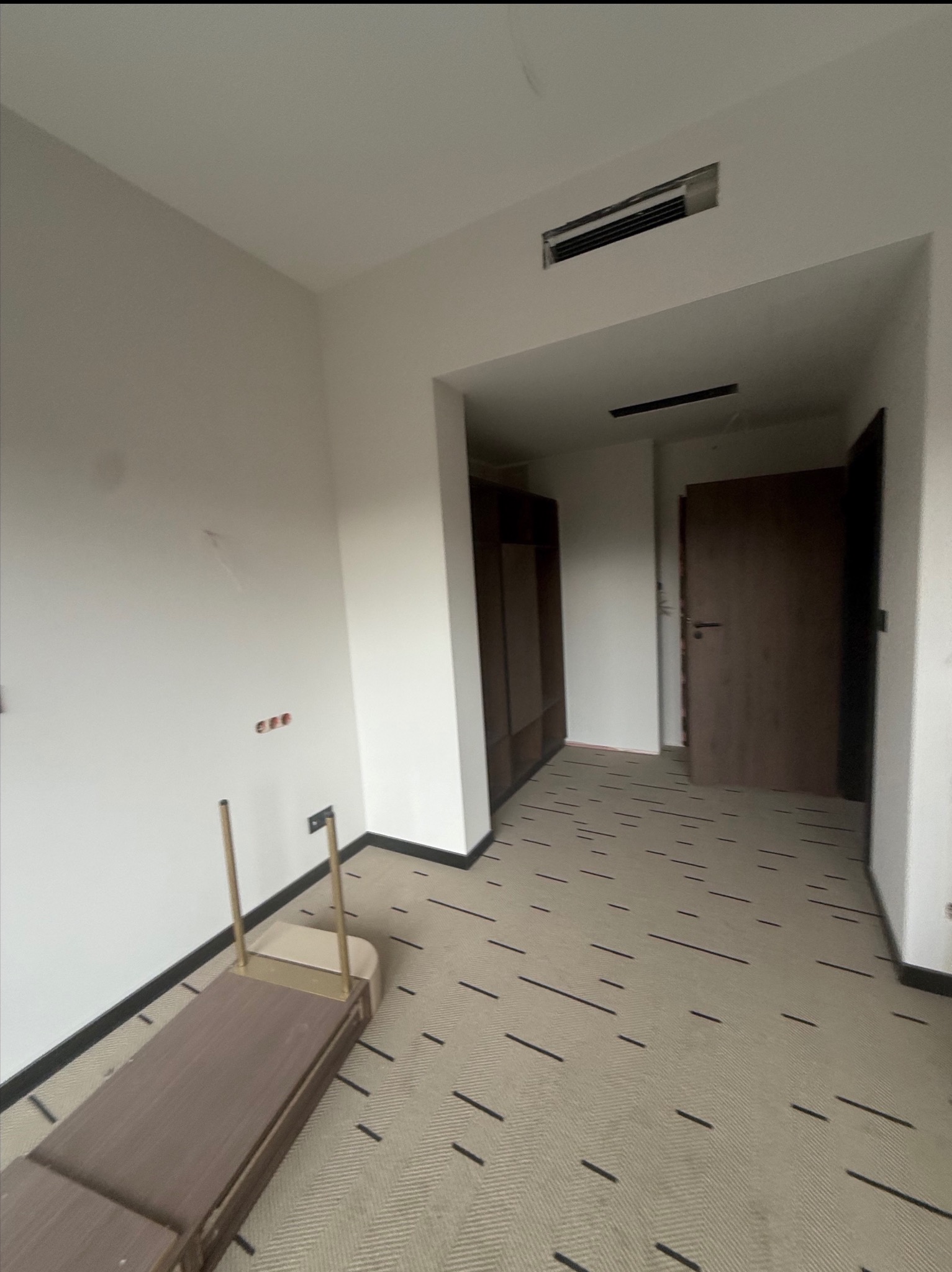 Wnętrze pokoju w trakcie remontu: białe ściany, podłoga w geometryczny wzór, widoczne gniazdka elektryczne i fragmenty mebli. Otwarta przestrzeń z drzwiami.