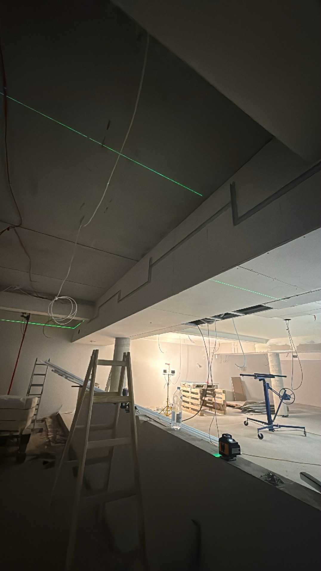 Montaż płyt gipsowo-kartonowych na suficie, widoczny laser krzyżowy, drabina i podnośnik. Prace wykończeniowe w surowym wnętrzu z paletami w tle.