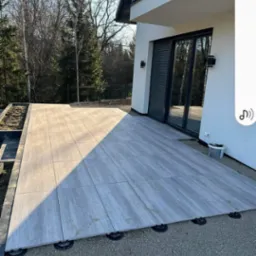 Nowoczesny taras z szarych płyt ułożonych na regulowanych wspornikach, otoczony żwirem, przylegający do jasnego budynku z dużymi oknami i ciemnymi żaluzjami.