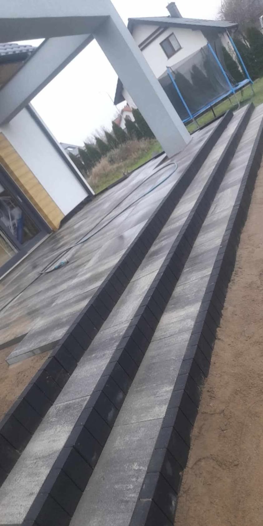Schody z kostki brukowej prowadzące do nowoczesnego domu z szarą elewacją i widocznym trampoliną w tle. Kąt ujęcia podkreśla trójwymiarowość konstrukcji.