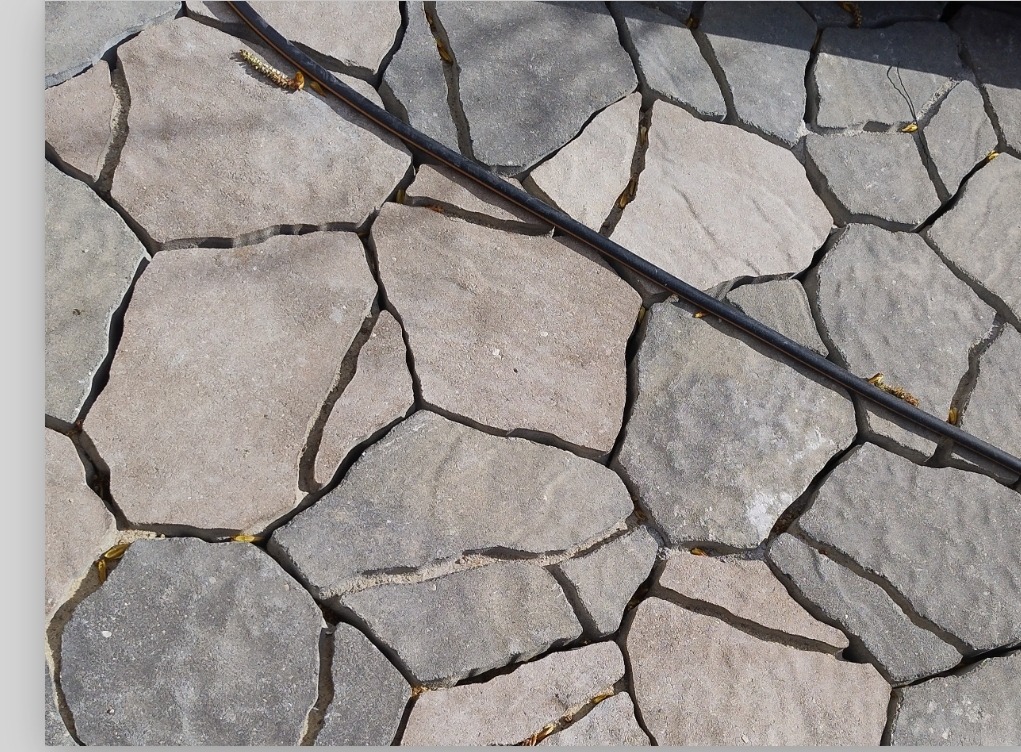 Nieregularna kostka brukowa w odcieniach szarości i beżu, ułożona na zewnątrz, z czarnym przewodem biegnącym po jej powierzchni. Widok z góry.