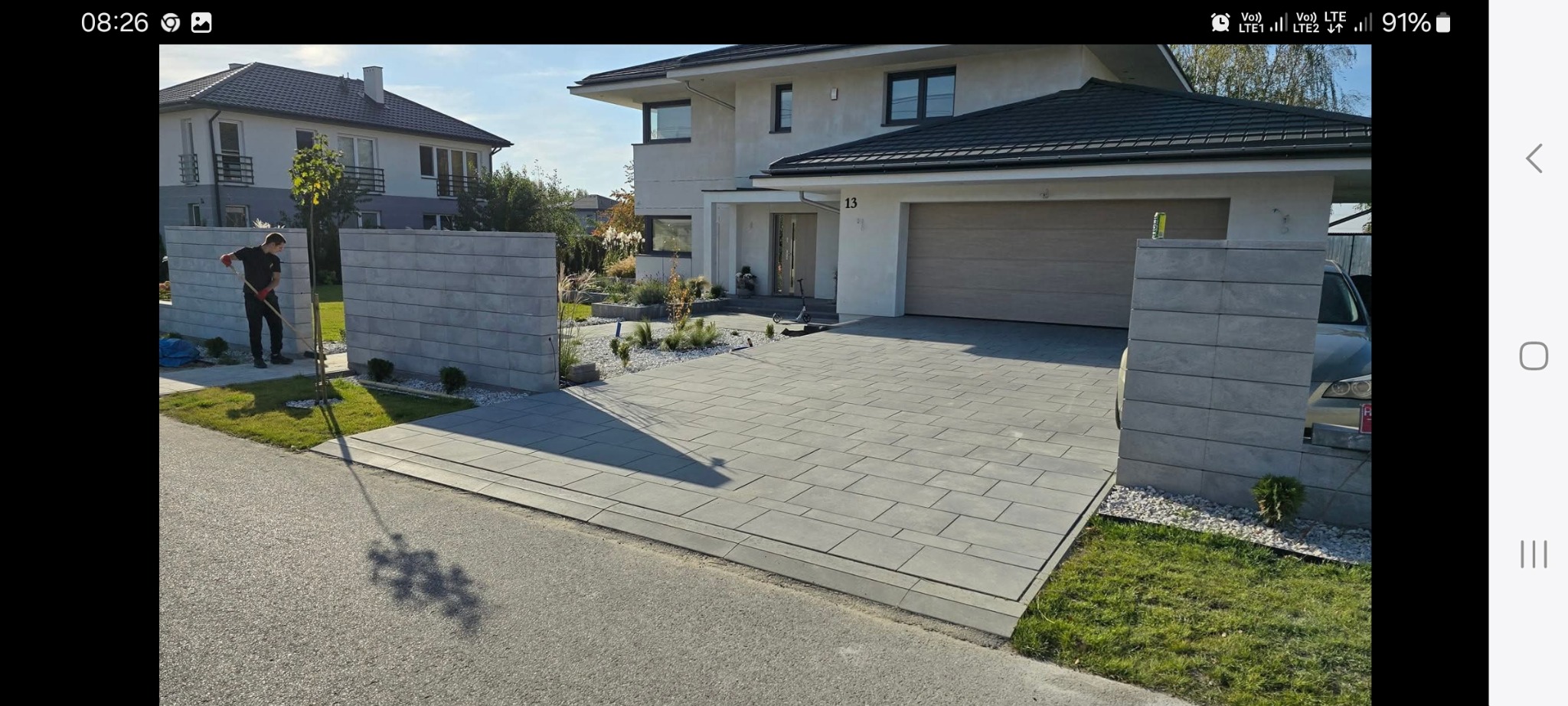 Nowoczesny podjazd z szarej kostki brukowej prowadzący do domu z garażem. Widoczne słupki ogrodzeniowe z tego samego materiału i pracownik w trakcie zamiatania.