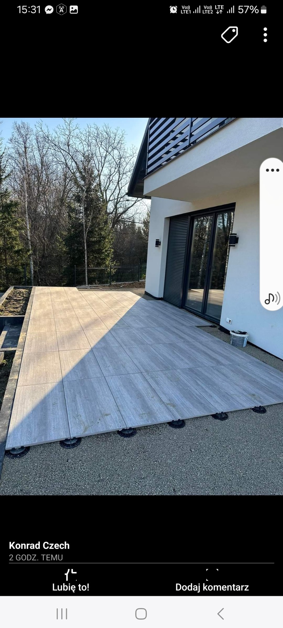 Nowoczesny taras z szarych płyt ułożonych na regulowanych wspornikach, otoczony żwirem, przylegający do jasnego budynku z dużymi oknami i ciemnymi żaluzjami.