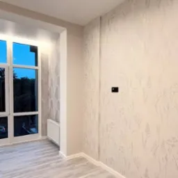 Jasne wnętrze z parkietem i ścianą w delikatne, roślinne wzory. Widoczne okno z widokiem na zieleń. Minimalistyczny styl wykończenia, czysty kadr.