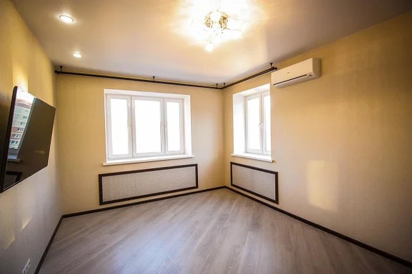 Jasne wnętrze pokoju po remoncie: panele podłogowe, okna z białymi ramami, ściany w odcieniach beżu, grzejniki za ozdobnymi kratkami, klimatyzacja, telewizor na ścianie.