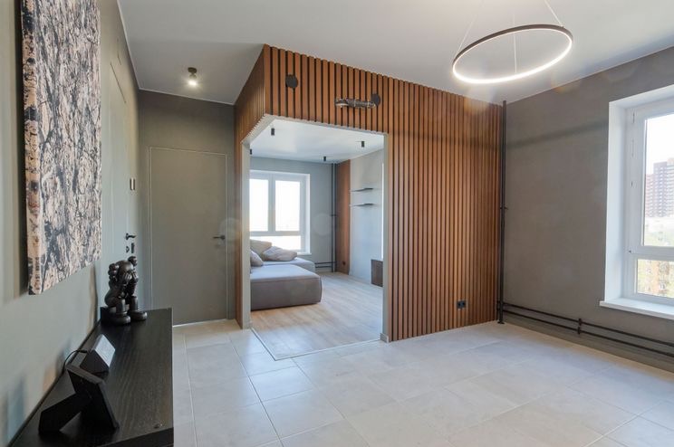 Nowoczesne wykończenie wnętrza: szare ściany, drewniana ścianka działowa z lamelami, okrągła lampa sufitowa i minimalistyczne dekoracje w jasnym, przestronnym pokoju.