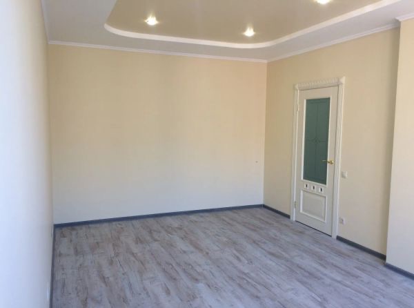 Jasne wnętrze pokoju z panelami podłogowymi, kremowymi ścianami i białymi drzwiami z przeszkleniami. Sufit podwieszany z oświetleniem punktowym.