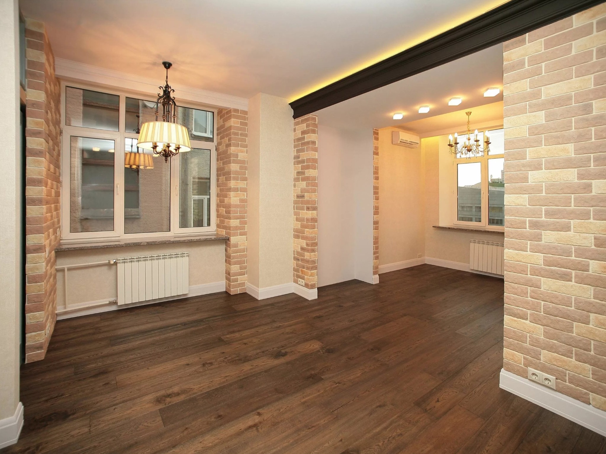 Wykończone wnętrze z ceglaną ścianą, drewnianą podłogą i oświetleniem sufitowym. Widoczne okna z grzejnikami i dekoracyjne lampy.