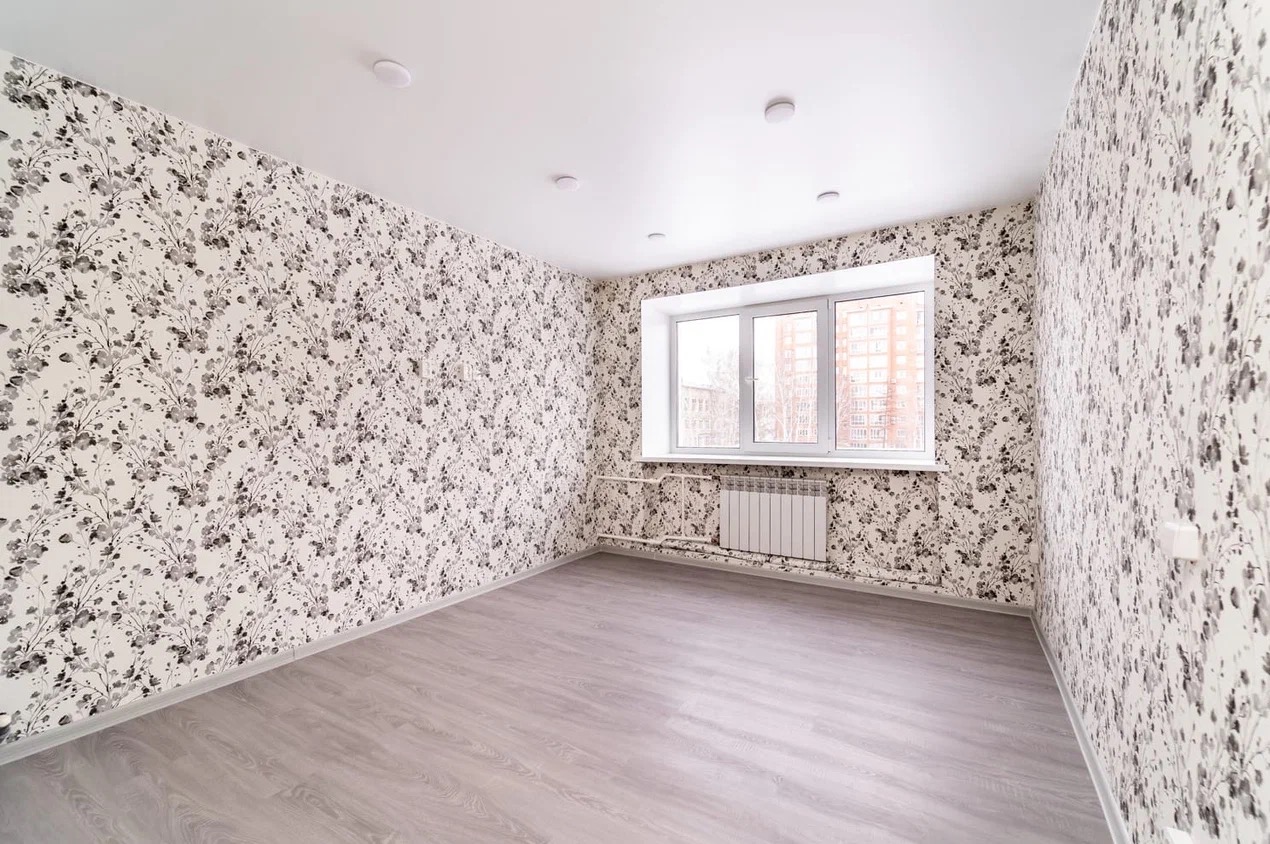 Pusty pokój po remoncie: ściany w kwiatowy wzór, jasna podłoga, białe okno z grzejnikiem. Widok na budynki za oknem. Nowoczesne oświetlenie sufitowe.