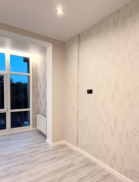 Jasne wnętrze z parkietem i ścianą w delikatne, roślinne wzory. Widoczne okno z widokiem na zieleń. Minimalistyczny styl wykończenia, czysty kadr.