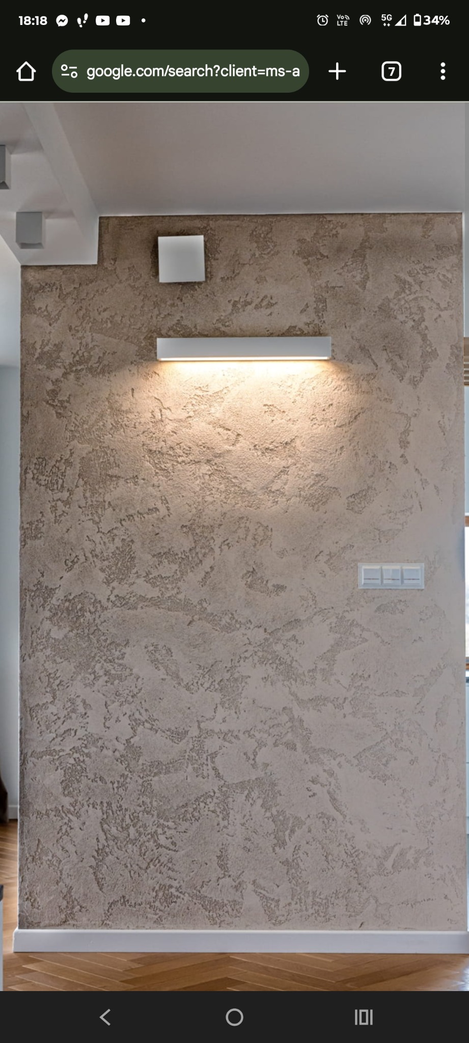 Dekoracyjna ściana z efektem trawertynu w odcieniach beżu, oświetlona podłużną lampą LED. Widoczne włączniki światła i fragment drewnianej podłogi.