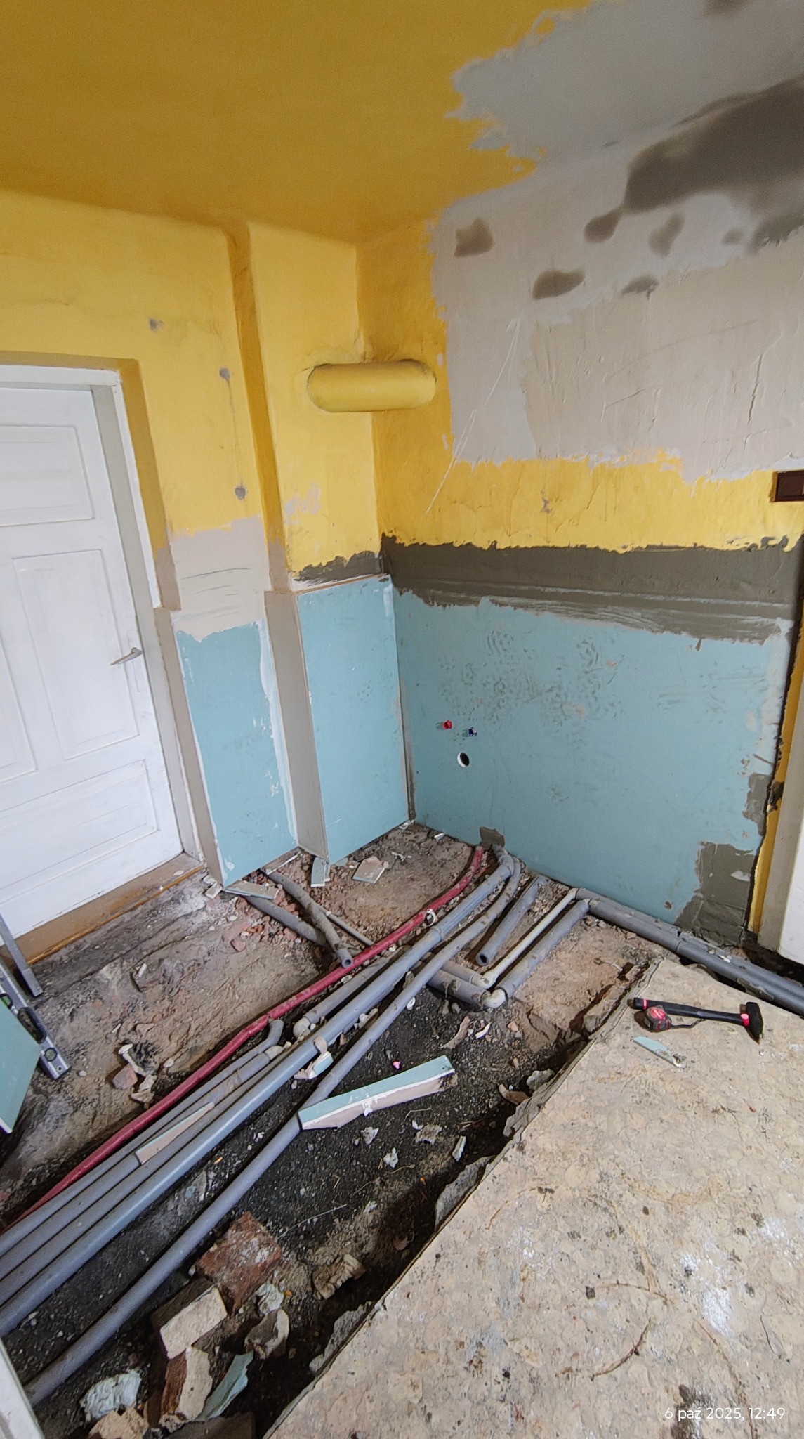 Żółto-szara łazienka w trakcie prac: ściany z fragmentami surowego tynku, rury na podłodze, płyty gipsowe, przygotowanie do dalszych prac wykończeniowych.