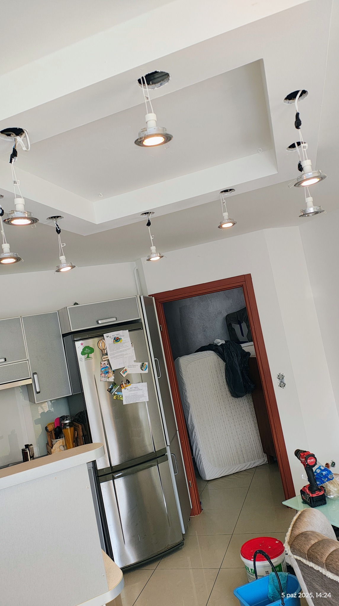 Kuchnia w trakcie montażu oświetlenia sufitowego: kilka lamp wpuszczanych, przewody widoczne. W tle lodówka, drzwi do pokoju z materacem i narzędzia pracy.