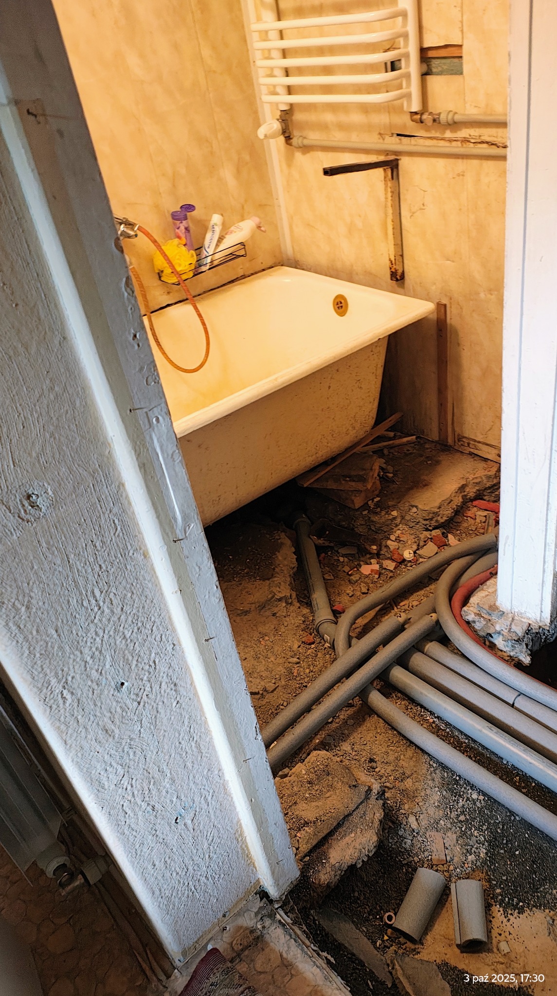 Łazienka w trakcie demontażu: widoczna wanna, odsłonięte rury i gruz. Fragment ościeżnicy po lewej stronie kadru.